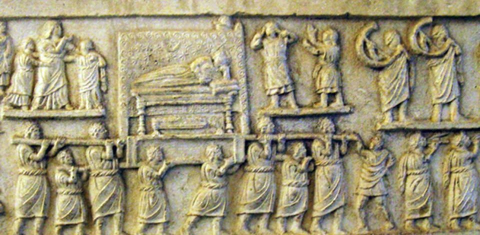 Ancient Roman stone relief depicting a funeral procession. Figures carry a decorated bier with a reclining body, accompanied by mourners, musicians playing curved horns, and others expressing grief with raised hands. The scene is carved in detailed, shallow relief.