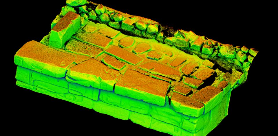 A colorized 3D scan of a stone structure segment, showing rectangular and rounded stones arranged in a wall and floor pattern. The scan uses a heatmap-style color gradient—red, orange, yellow, and green—to indicate elevation or depth, set against a black background.